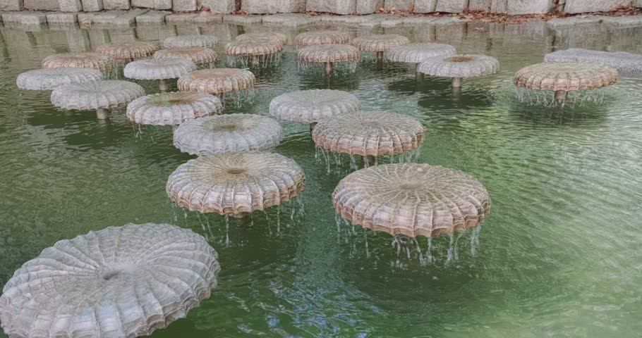 Close-up shot of unique stone mushroom-shaped fountain heads with water cascading down into a calm park pond in Munich, Germany, on a sunny day.