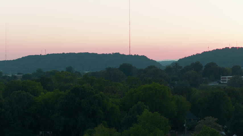 Appalachian foothills sunset above Knoxville, Tennessee: forested ridges layered into hazy distance under a pink and orange sky, summer canopy in the foreground.