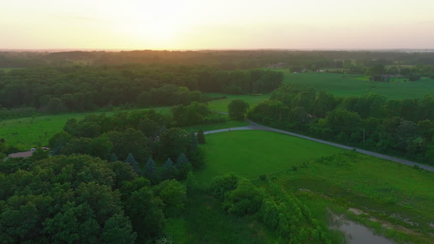 Aerial view of countryside at sunset near Racine, Wisconsin. Drone shot over forest, green fields, farmland and a country road at golden hour, hazy summer sky on the horizon.