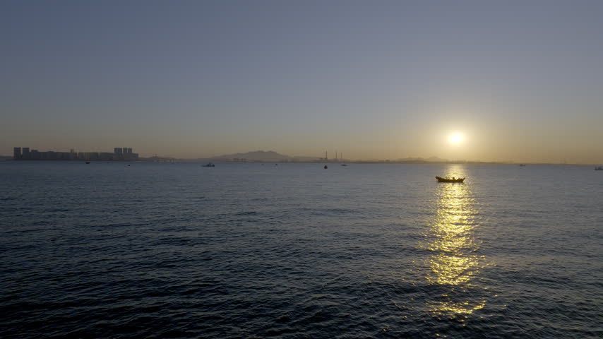 A serene seascape at sunset, featuring a small boat on calm water, golden sun reflection, and distant coastal structures under a clear sky.