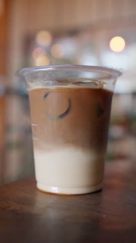 Plastic straw being inserted into an iced latte in a clear plastic cup, with visible milk layers and ice cubes, showcasing a refreshing cold coffee beverage and cafe lifestyle.