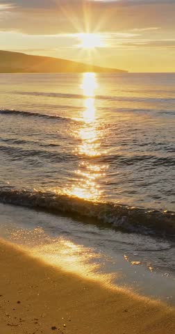 Cinematic footage of golden sun rising above horizon across ocean surface with soft waves rolling onto sandy beach.