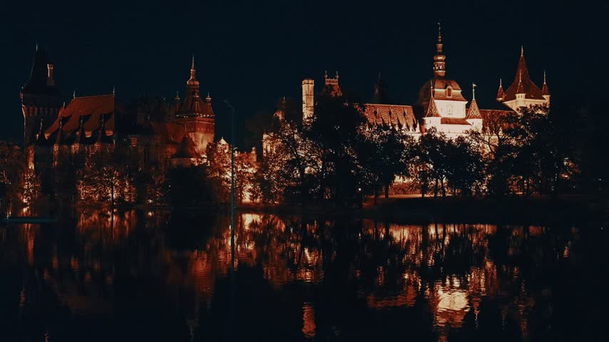 Cinematic night view of the illuminated Vajdahunyad Castle with golden light reflections shimmering across the calm lake surface in Budapest, Hungary.