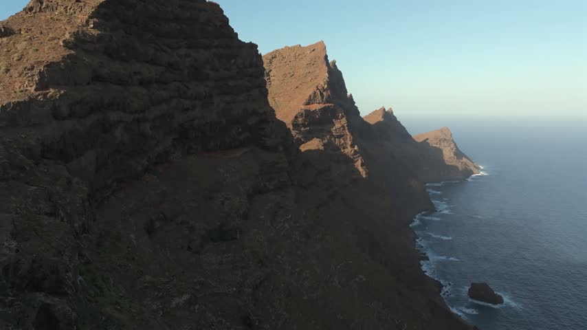 Golden hour aerial flight along towering dark volcanic sea cliffs on the west coast of Gran Canaria, Canary Islands, with the Atlantic far below