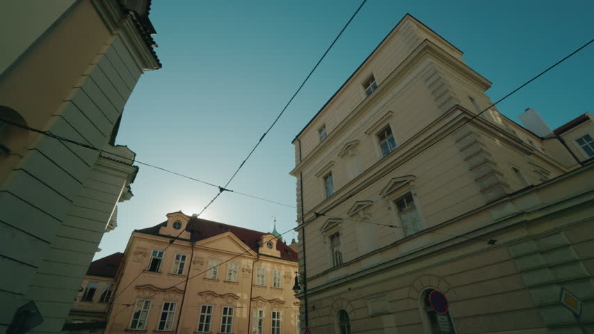 Sunlit historic facades and a church frame a narrow Prague street beneath a clear blue sky.