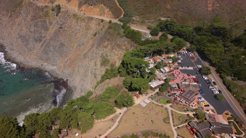 High oblique view of Ragged Point on Big Sur coastline, showing steep layered cliffs, turquoise water, white surf, offshore rocks, resort buildings, road access, and bright coastal landscape