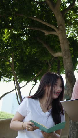 Young woman in white t-shirt reads notebook outdoors under trees during bright daylight.