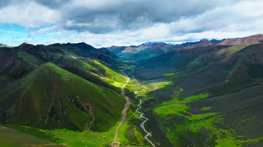 Elevated view of a winding stream and dirt road crossing a sunlit mountain valley beneath dramatic clouds and distant snowy peaks.