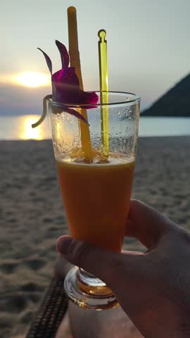 First person view of a man lying on a lounge chair on the sand, holding a tall glass of orange juice garnished with flowers and straws. The sun is setting over the ocean.