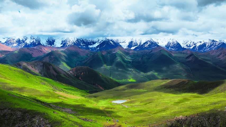 Sunlit alpine grassland rolls toward a small pond beneath a distant snowcapped range, with dramatic clouds casting shadows across the valley.