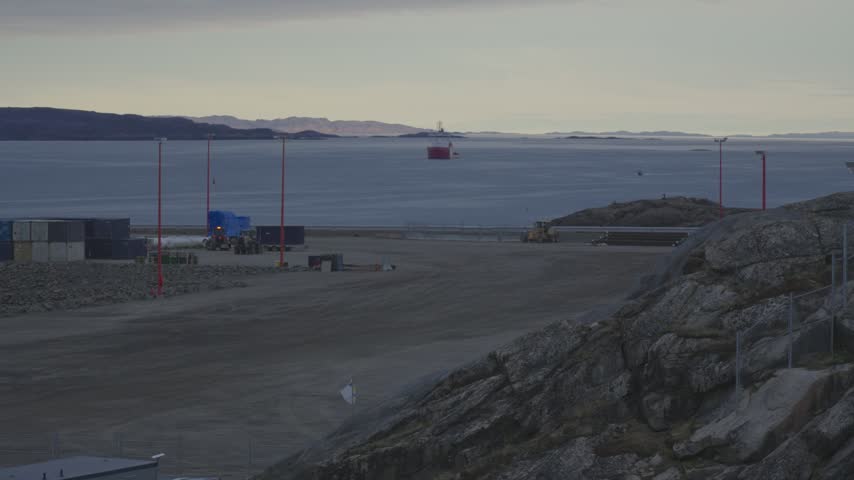 Wide ground level view across rocky port staging ground toward Frobisher Bay shipping with vessel Captain Molly Kool anchored in distance
