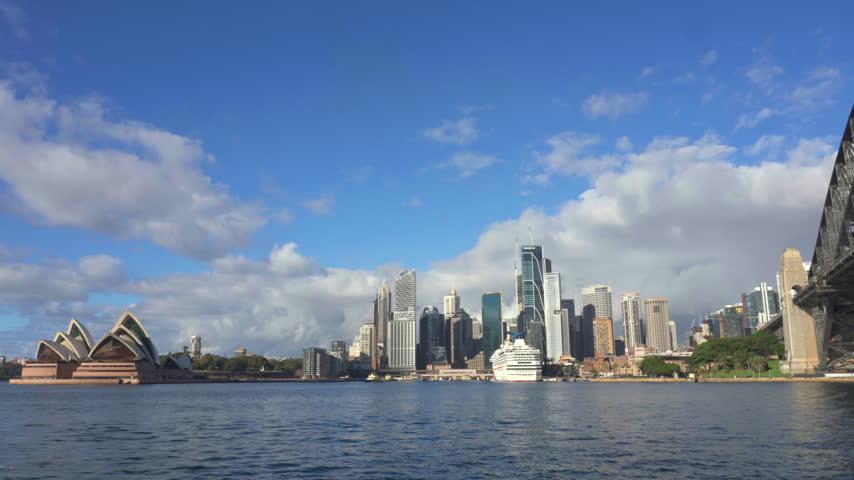 Wide shot across Sydney Harbour featuring the Opera House, CBD skyline, cruise ship and edge of the Harbour Bridge beneath a bright blue sky. Sydney, New South Wales, Australia.