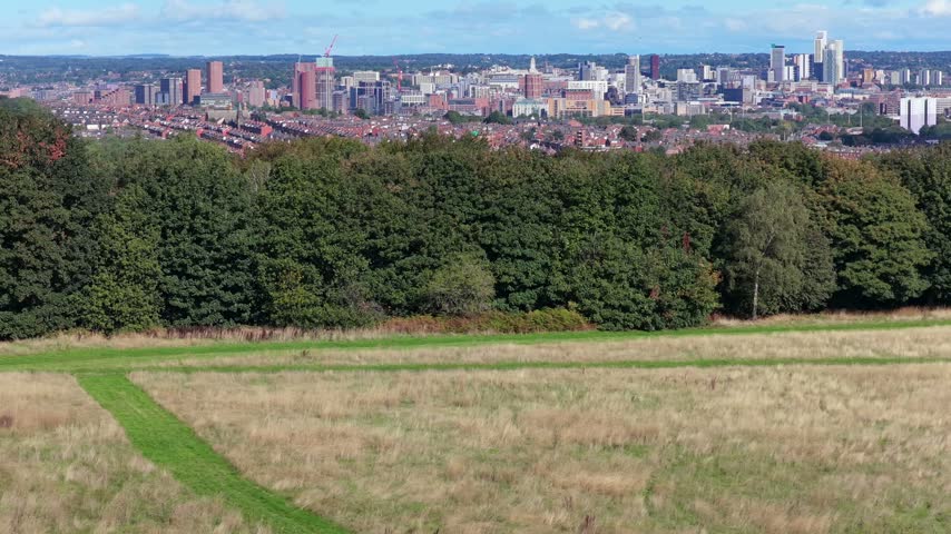 Elevating aerial video looking north across the open grassy slopes and paths of the aeromodellers flying field at Middleton Park, leading over a belt of woodland trees toward suburban housing and the distant Leeds city centre skyline under a bright, partly cloudy sky