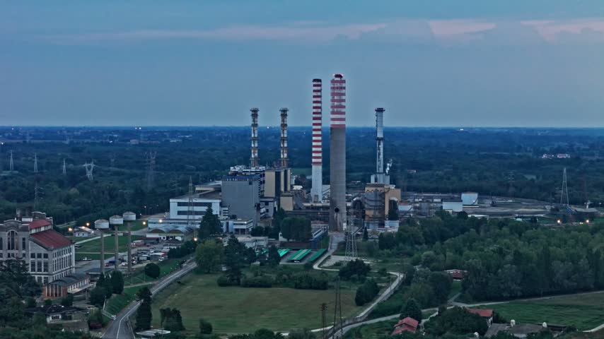 Industrial landscape showing an electric power plant with tall chimneys at dusk. A commuter train is moving across the foreground