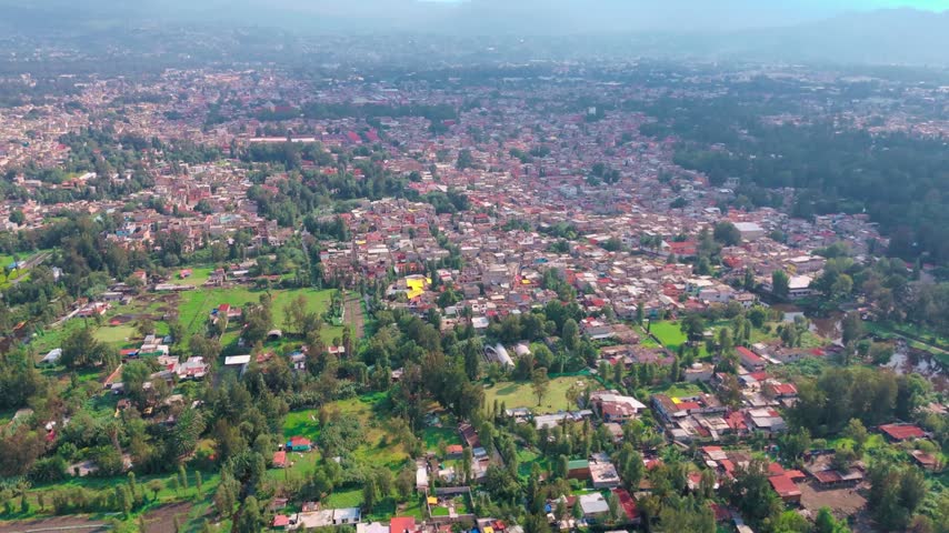 Aerial view of the rural chinampa landscape meeting dense urban housing in Xochimilco, Mexico City, with green agricultural areas and trees throughout the city.