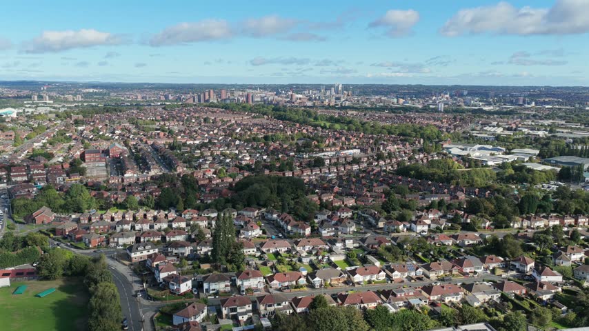 Aerial video looking north from Middleton across Cockburn School grass playing fields and artificial sports pitches, capturing suburban Beeston housing and the distant Leeds city skyline under a clear morning sky.