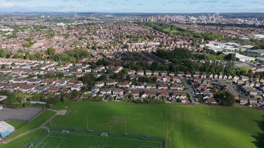 Aerial video looking north from Middleton across Cockburn School grass playing fields and artificial sports pitches, capturing suburban Beeston housing and the distant Leeds city skyline under a clear morning sky.