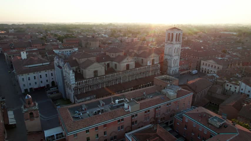 A drone orbits the marble facade, arcaded loggia and brick campanile of Ferrara Cathedral while low sunrise light flares over the terracotta rooftops of the old town. The Cattedrale di San Giorgio stands in the historic centre of Ferrara, a Renaissance city in Emilia-Romagna listed as UNESCO World Heritage.