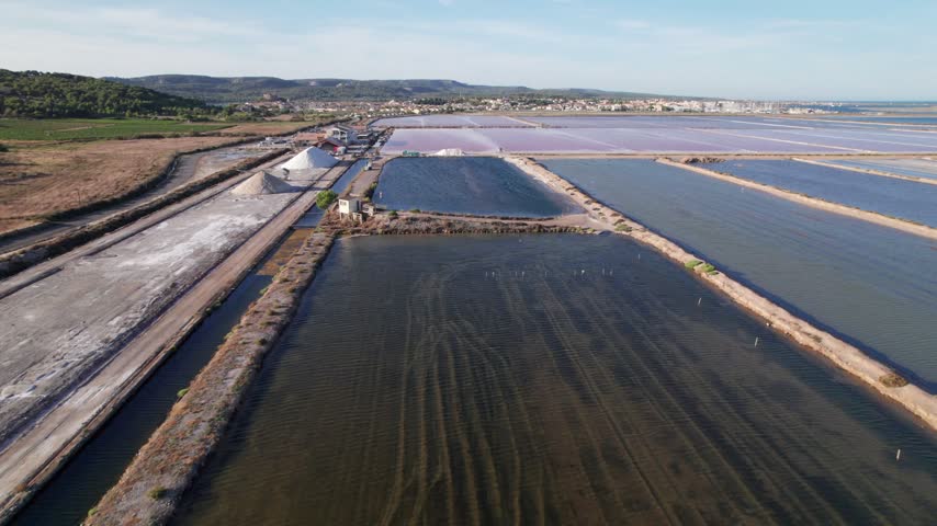 A drone flies forward low over a dark brine basin towards white salt mounds and bright pink evaporation ponds, with the salt works buildings and the town of Gruissan on the horizon at sunset. Le Salin de l