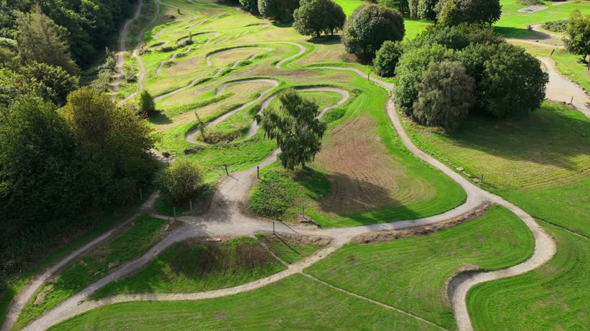 Aerial video looking down across the winding dirt tracks, berms, and grassy terrain of the Middleton Park bike trails and pump track facility, bordered by lush green parkland trees under bright daytime sunlight.