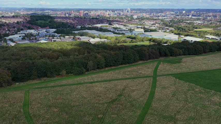 Aerial video showcasing the large open flying field and criss-crossing mown runways of the aeromodellers grounds at Middleton Park, enveloped by dense woodland canopies, looking north toward South Leeds suburbs and the distant city centre skyline under a bright, cloud-filled sky.