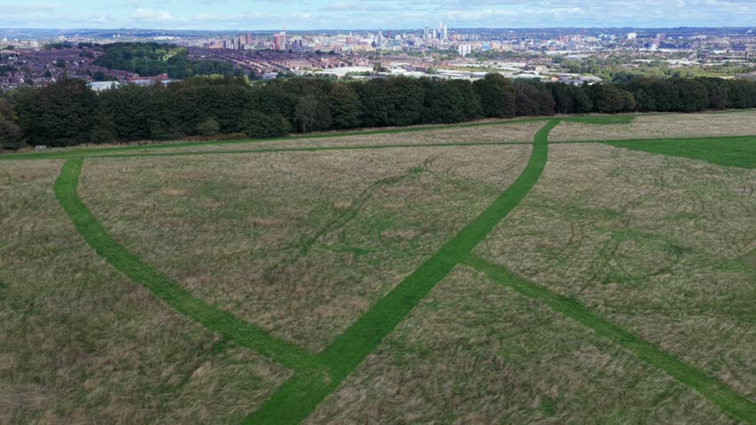 Aerial video showcasing the large open flying field and criss-crossing mown runways of the aeromodellers grounds at Middleton Park, enveloped by dense woodland canopies, looking north toward South Leeds suburbs and the distant city centre skyline under a bright, cloud-filled sky.