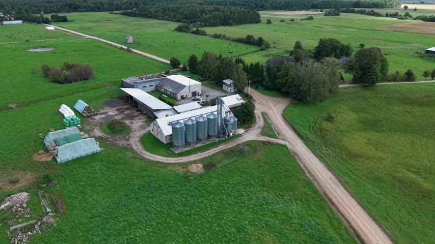 Aerial footage captures a rural farm complex with silos, barns, and winding dirt roads surrounded by lush green fields on a clear day, providing a broad view of agricultural land.