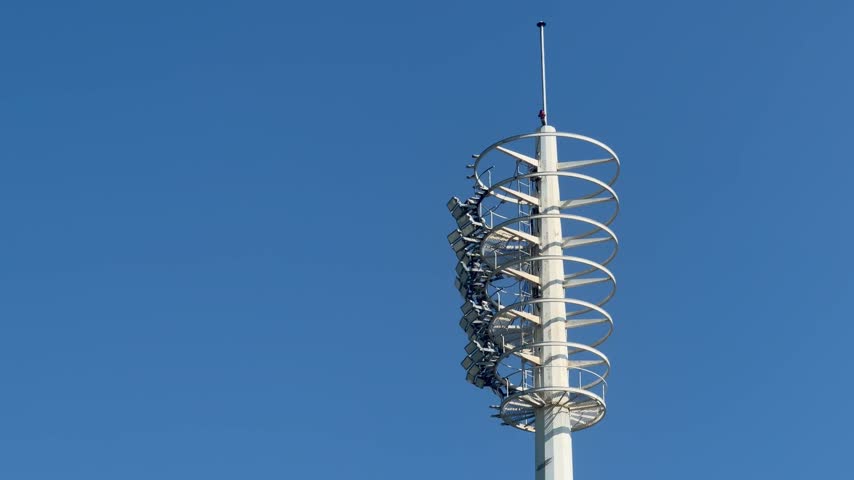 A tall tiered high-mast floodlight tower with multiple rings of stadium lighting fixtures, set against a clear blue sky