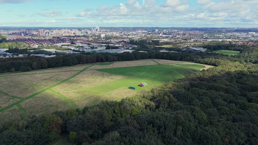 Aerial video showcasing the large open flying field and criss-crossing mown runways of the aeromodellers grounds at Middleton Park, enveloped by dense woodland canopies, looking north toward South Leeds suburbs and the distant city centre skyline under a bright, cloud-filled sky.