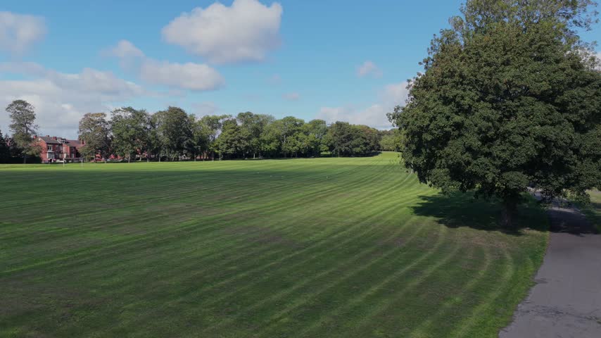 Aerial video perspective overlooking the expansive mown grass football pitches, tree-lined walking avenues, and recreation grounds of Cross Flatts Park in Beeston, bordering red-brick Victorian terraced housing under a bright, partly cloudy sky.