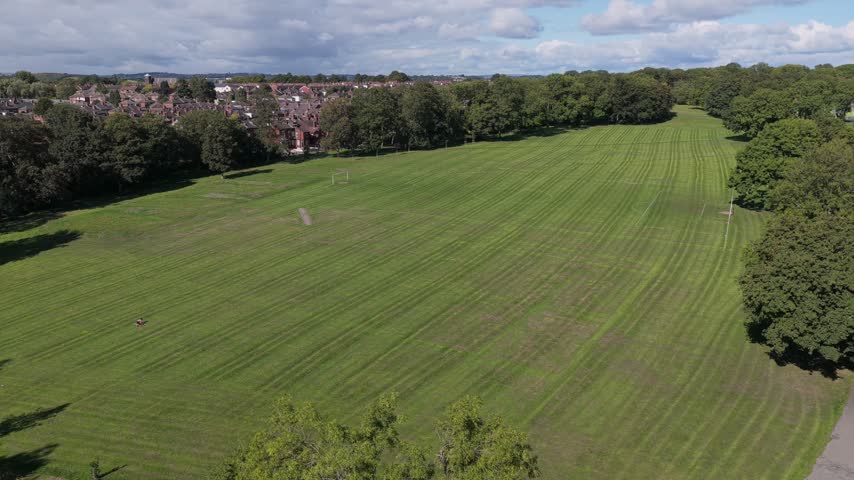 Aerial video capturing the sprawling green lawns, recreational grounds, and mature tree lines of Cross Flatts Park bordering the dense Victorian terraced housing of Beeston, looking north towards the Leeds city centre skyline under a bright, cloud-filled blue sky.