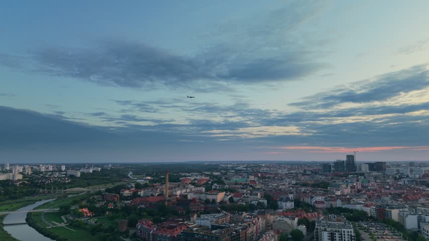 Stunning aerial panorama of Poznan in Poland during a colorful sunset, showing the cityscape with historic architecture, modern buildings, and an airplane flying across the beautiful evening sky