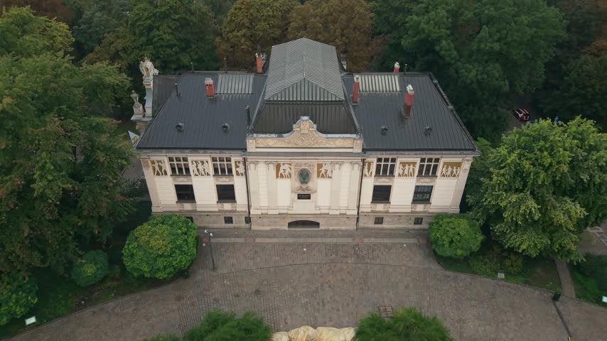 Aerial view descending towards the elegant facade of the Palace of Art in Krakow, Poland, showcasing its detailed neoclassical architecture and the beautiful fountain appearing in the foreground