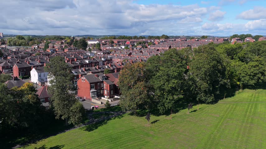 Aerial video perspective capturing the green slopes and tree-lined border of Cross Flatts Park running alongside the extensive red-brick Victorian terraced streets of Beeston under a bright blue sky filled with cumulus clouds.