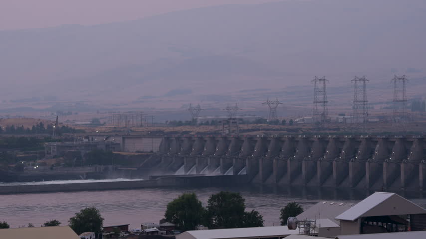 View of The Dalles Dam on the Columbia River with concrete spillways, power transmission towers, and rolling hills in the background in The Dalles, Oregon, United States.