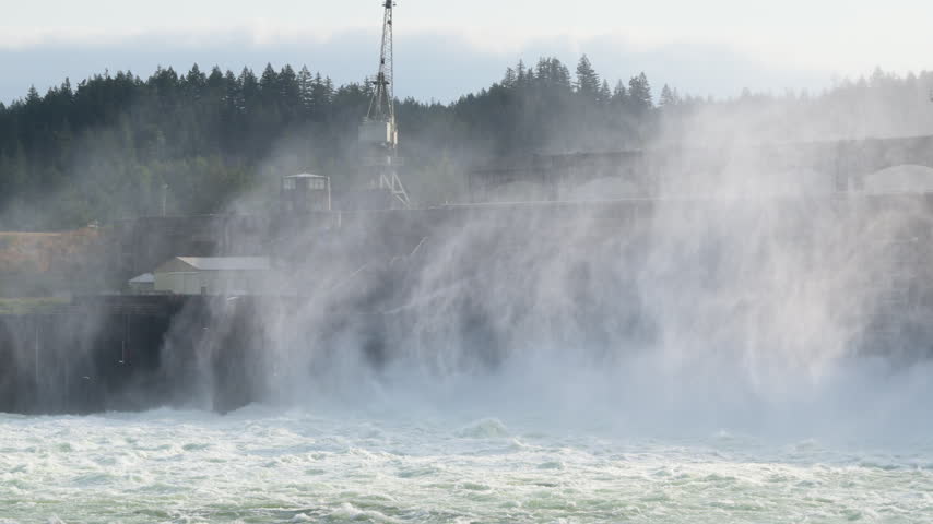 Turbulent water surging through Bonneville Dam on the Columbia River in Oregon, creating towering spray and mist around the hydroelectric facility. Slow motion panning shot