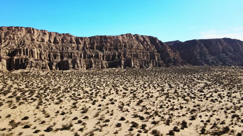 Sparse shrubs dot a sandy desert plain beneath a tall rocky ridge in Catamarca, Argentina, as the drone flies steadily forward under clear blue skies.