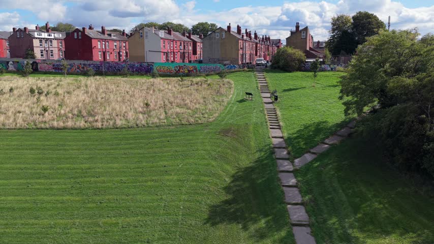 Aerial video showcasing the green lawn, wildflower meadow patch, and paved pathway of Holbeck Moor Park, bordering colourful mural walls and dense Victorian red-brick terraced housing in Beeston under a bright, cloud-filled sky.