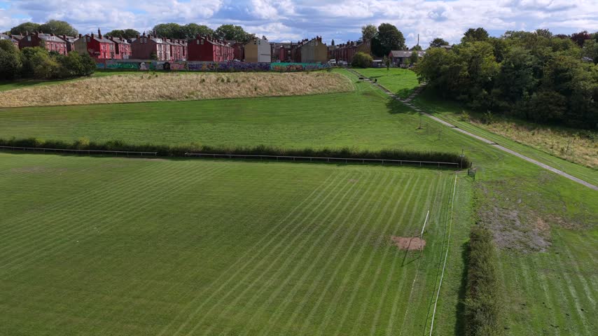 Aerial video showcasing the green lawn, wildflower meadow patch, and paved pathway of Holbeck Moor Park, bordering colourful mural walls and dense Victorian red-brick terraced housing in Beeston under a bright, cloud-filled sky.