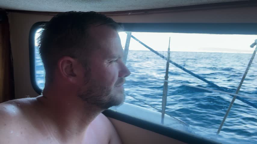 Man looking through a boat cabin window at the moving sea during a sailing journey.
