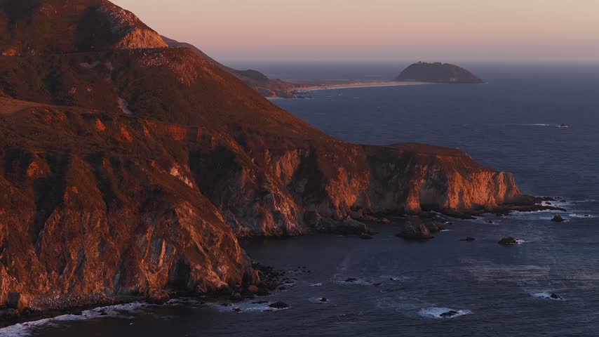 Aerial view gliding along the rugged rocky cliffs and Pacific Ocean surf near Big Sur, California, revealing Point Sur Lighthouse in the distance at sunset.