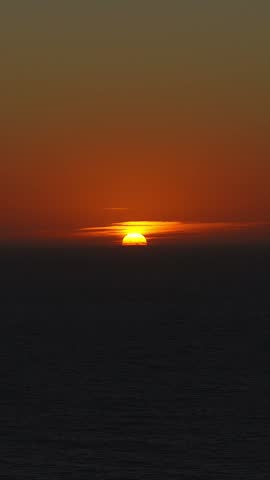 Golden sun sets behind low clouds across the dark ocean horizon near Big Sur California during dusk. The warm sky transitions to night over the quiet water.