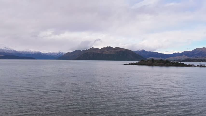 Wide aerial view of Ruby Island on Lake Wanaka with distant mountains and cloudy sky
