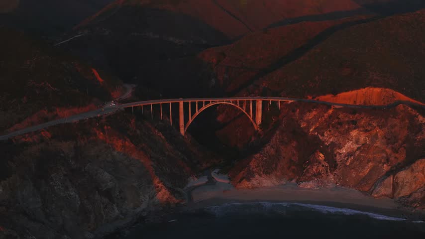 Aerial shot displays Bixby Creek Bridge in Big Sur California spanning a rugged canyon, with warm late afternoon light grazing rocky cliffs and ocean waves breaking along the beach below.