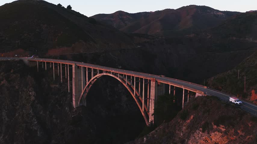 Aerial view tracks Bixby Creek Bridge spanning a deep canyon along Highway 1 in Big Sur, California, illuminated by warm sunset light along the mountainous Pacific coastline.