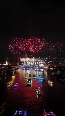 Colorful fireworks illuminating the night sky over a city river during a national holiday celebration in Bangkok, Thailand, with illuminated tourist boats watching the spectacular pyrotechnic show.