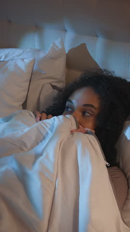 African American girl in bed pretending scary afraid covering duvet smiling laughing happy looking at camera girl female pretend frightened with nightmare darkness smile laugh at dark bedroom joke fun