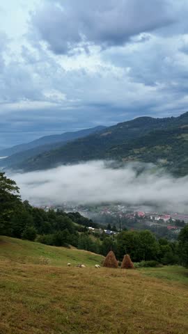Vertical shot of an idyllic mountain landscape on an overcast day. Scenic view of a rural valley covered in mist, green slopes, forest, and traditional haystacks under a cloudy sky.