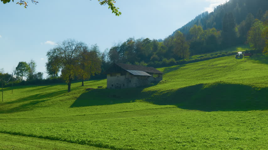 Alpine meadow rural barn on a rolling green hillside with a tractor working in the background. Handheld left to right pan shot of a traditional countryside landscape under a blue sky on a sunny day.
