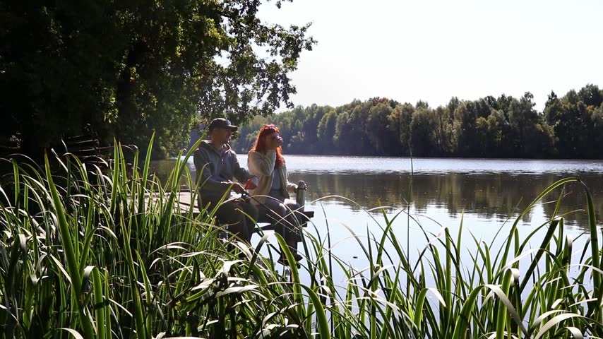 A man and a woman are sitting side by side on a wooden pier by the lake. Fishing together, relaxing together in nature and peaceful leisure by the water.
